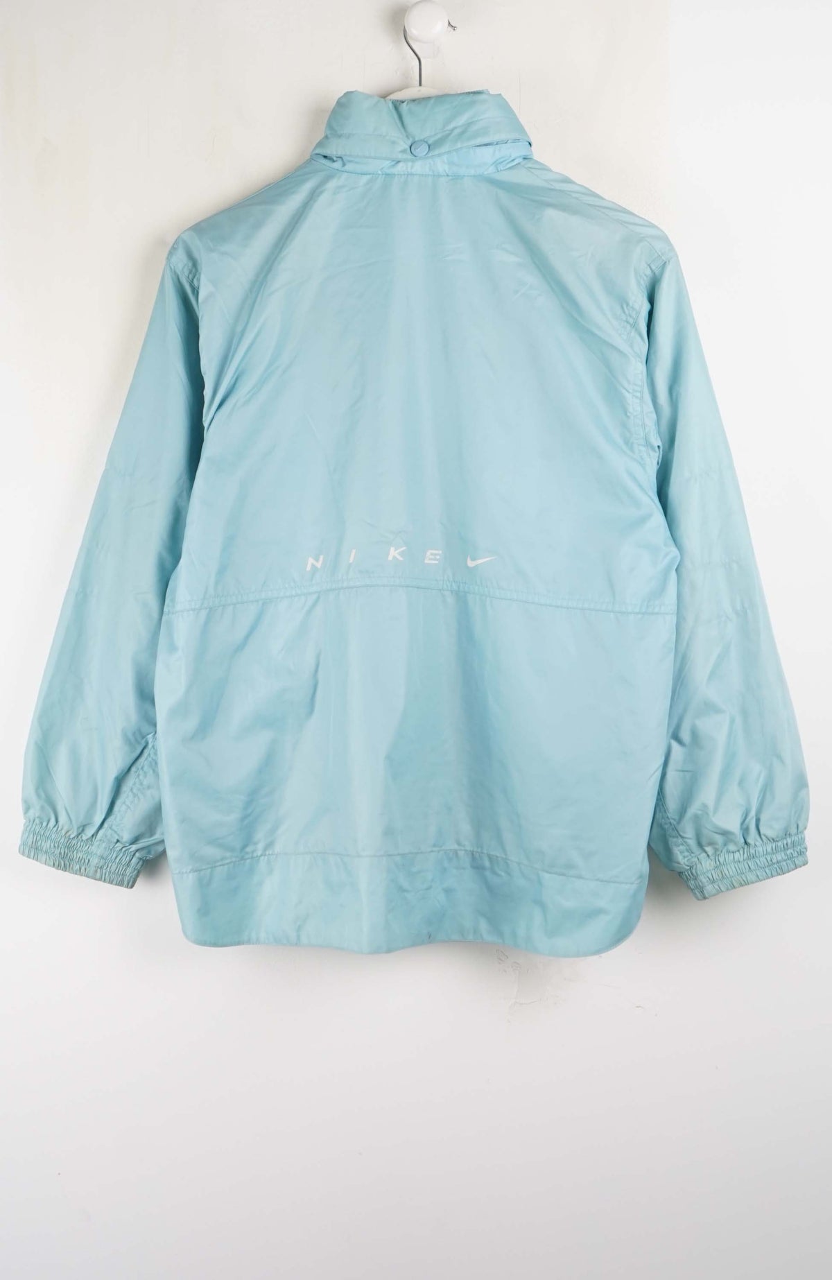 A light blue Nike windbreaker jacket hanging on a white hanger against a plain white wall, showing the back view with a small white Nike logo.