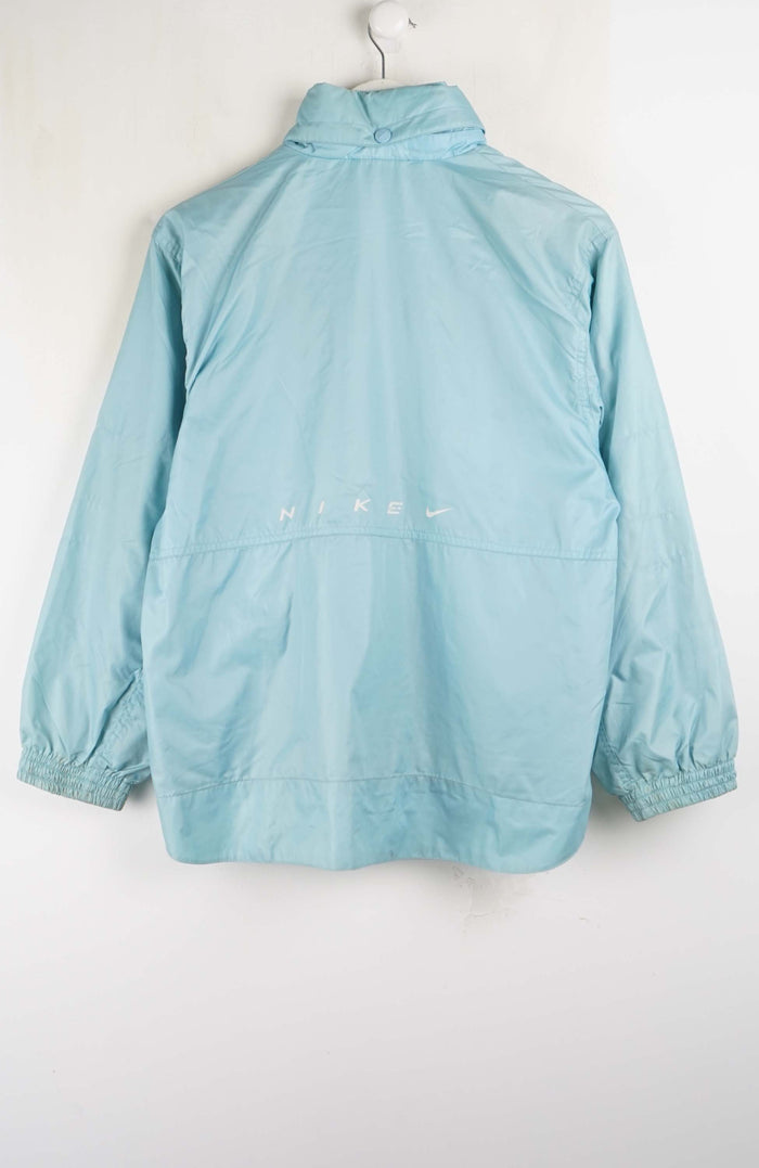 A light blue Nike windbreaker jacket hanging on a white hanger against a plain white wall, showing the back view with a small white Nike logo.
