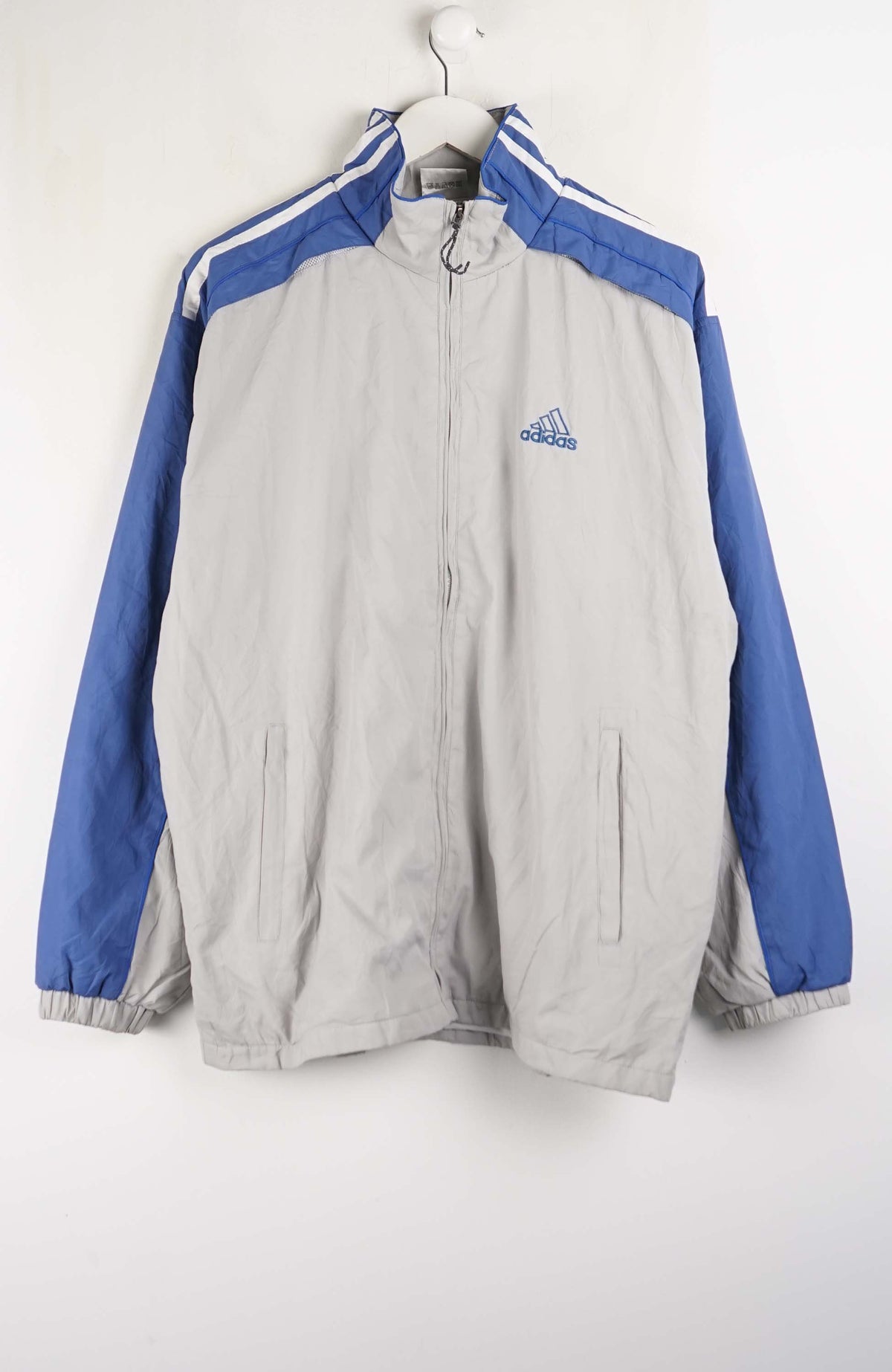 An Adidas windbreaker jacket in light gray and blue, hanging on a white hanger against a white wall. The jacket features a light gray body, royal blue sleeves, and white stripes on the shoulders, with the blue Adidas logo on the left chest.