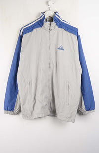 An Adidas windbreaker jacket in light gray and blue, hanging on a white hanger against a white wall. The jacket features a light gray body, royal blue sleeves, and white stripes on the shoulders, with the blue Adidas logo on the left chest.